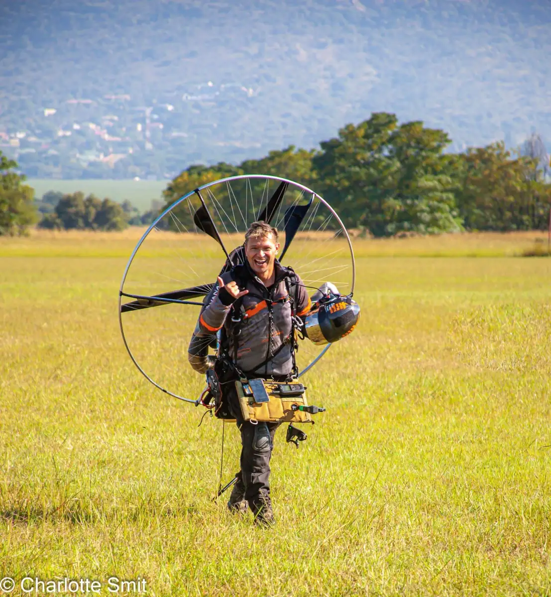 Eugene Cussons smiling and giving a shaka sign while carrying his paramotor across a field
