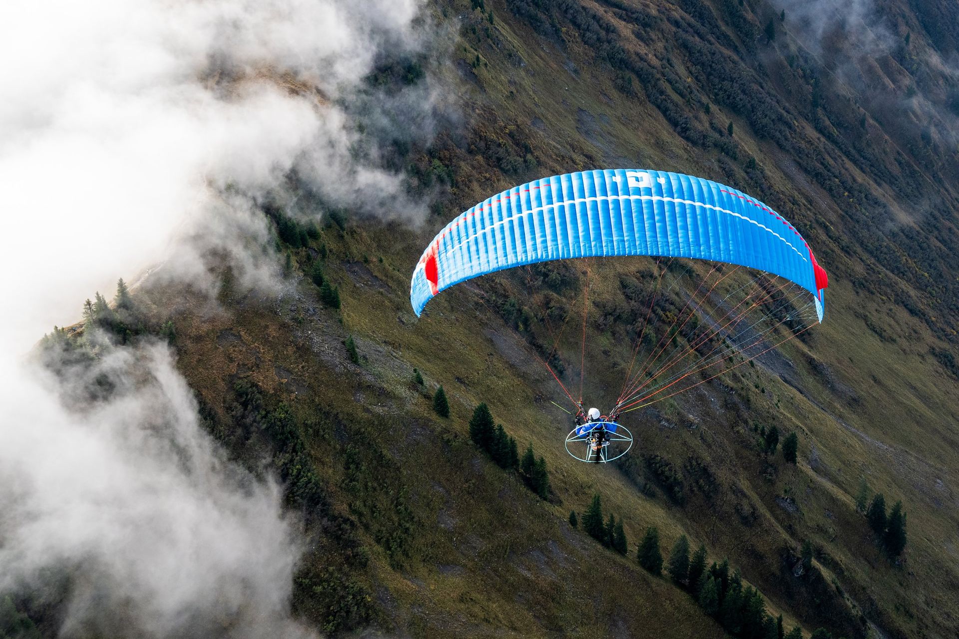 A blue paramotor wing flying along a forested mountain slope with cloud