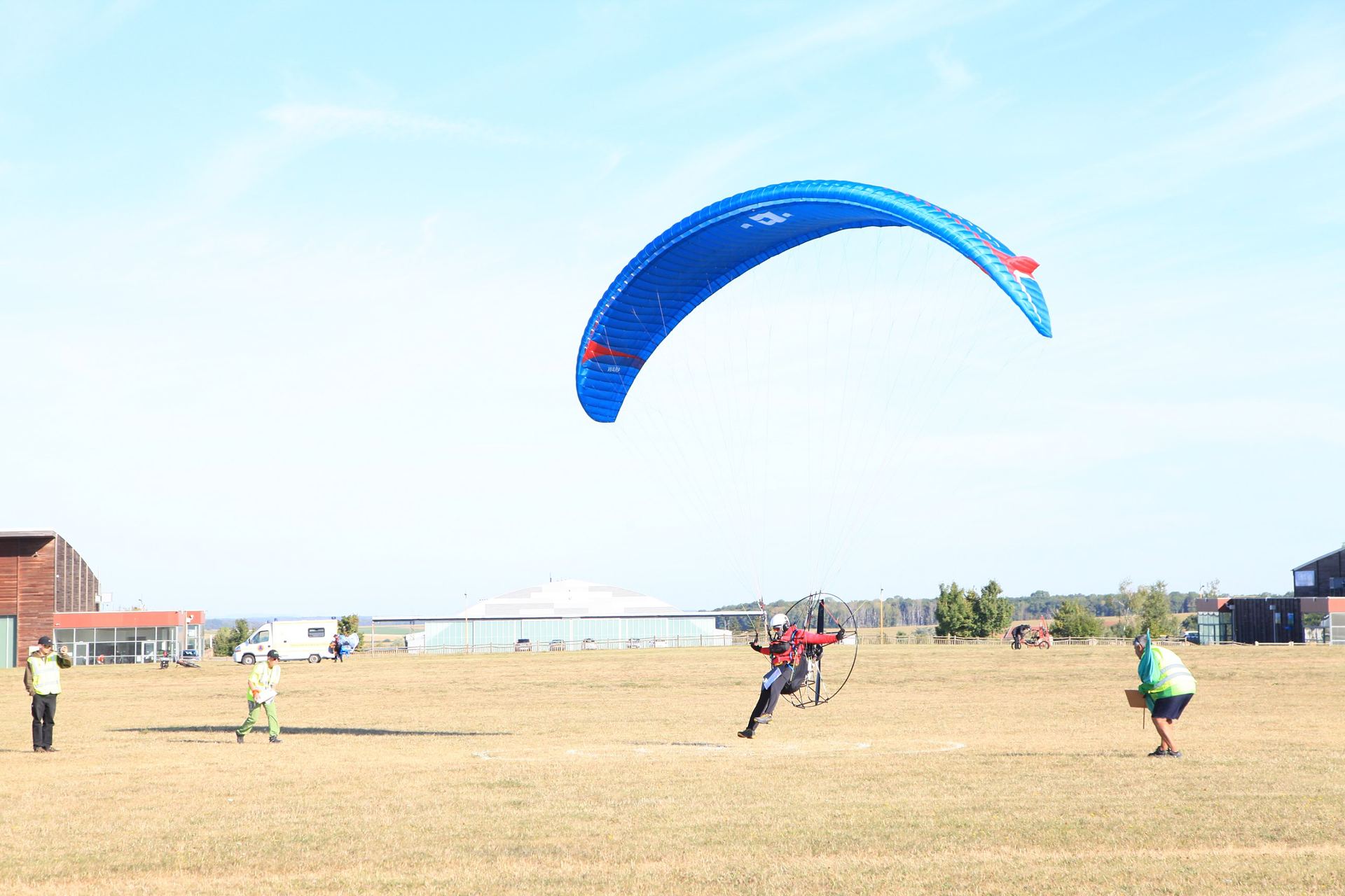 A pilot on a paramotor taking off under a blue wing on an airfield with helpers nearby