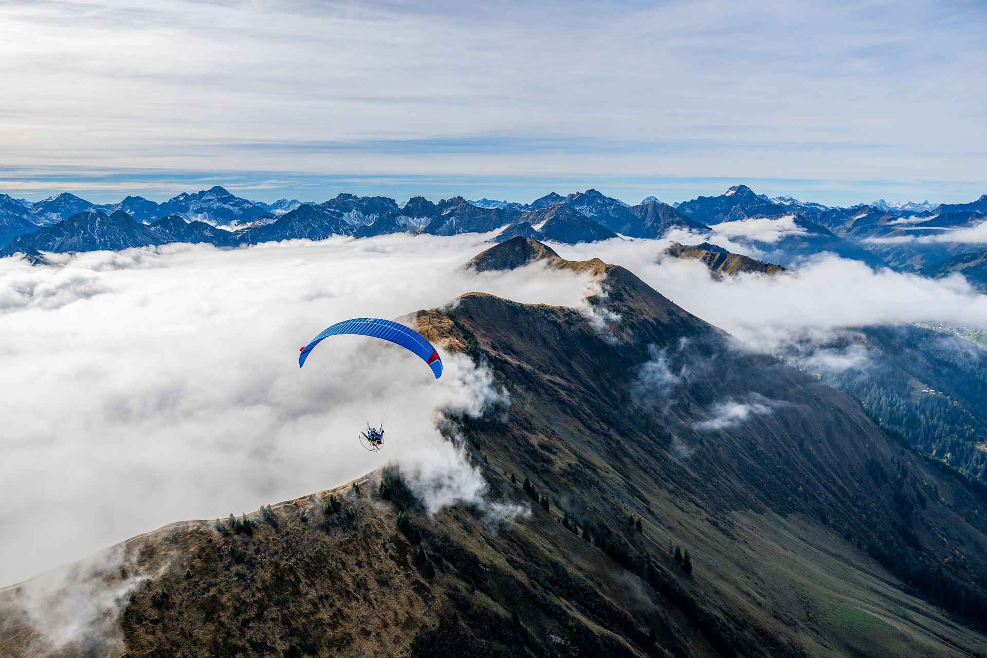 A paramotor wing flying along a mountain ridge above clouds