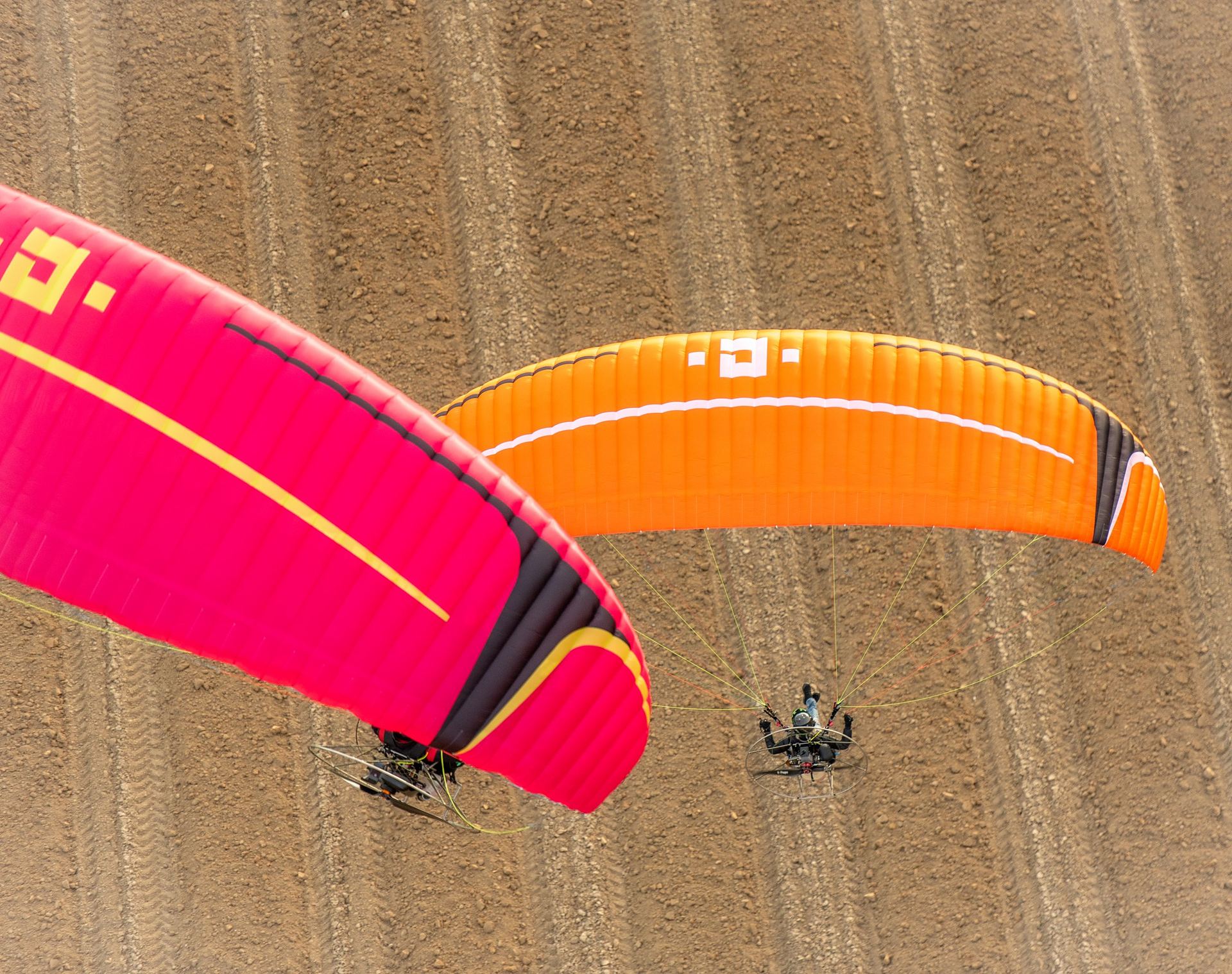 Pink and orange paramotor wings flying low over ploughed fields, seen from above
