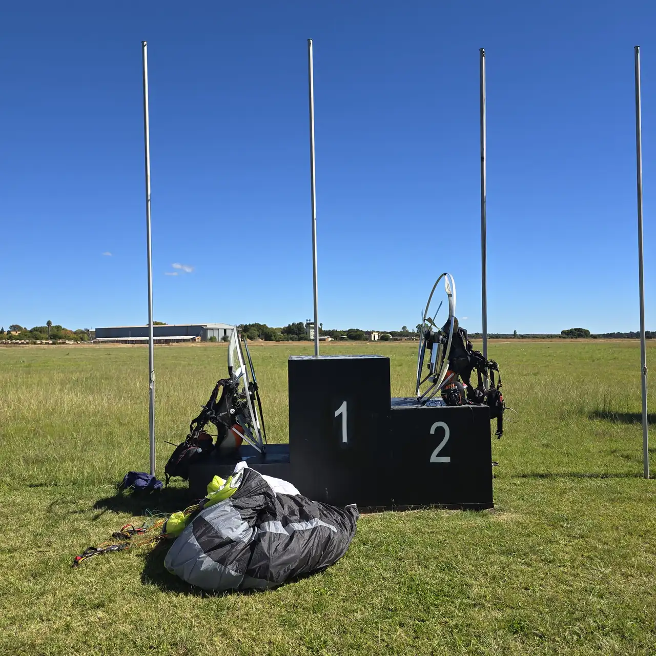 Two paramotors and a wing at the start field on Day 1, beside a winners podium and flagpoles