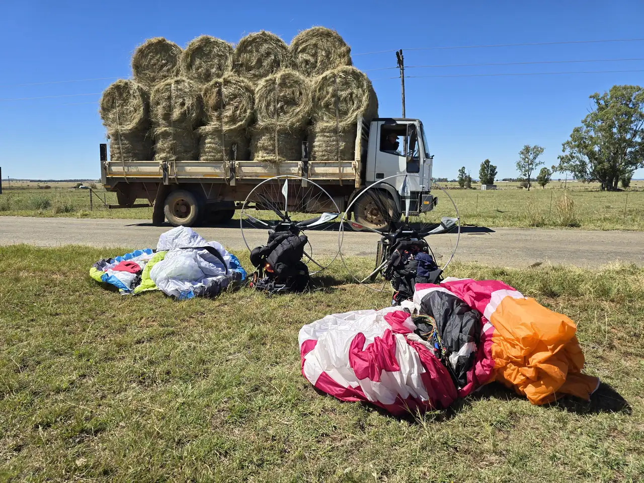 Two paramotors and wings on the grass beside a road, with a truck loaded with hay bales behind, after a landing on Day 1