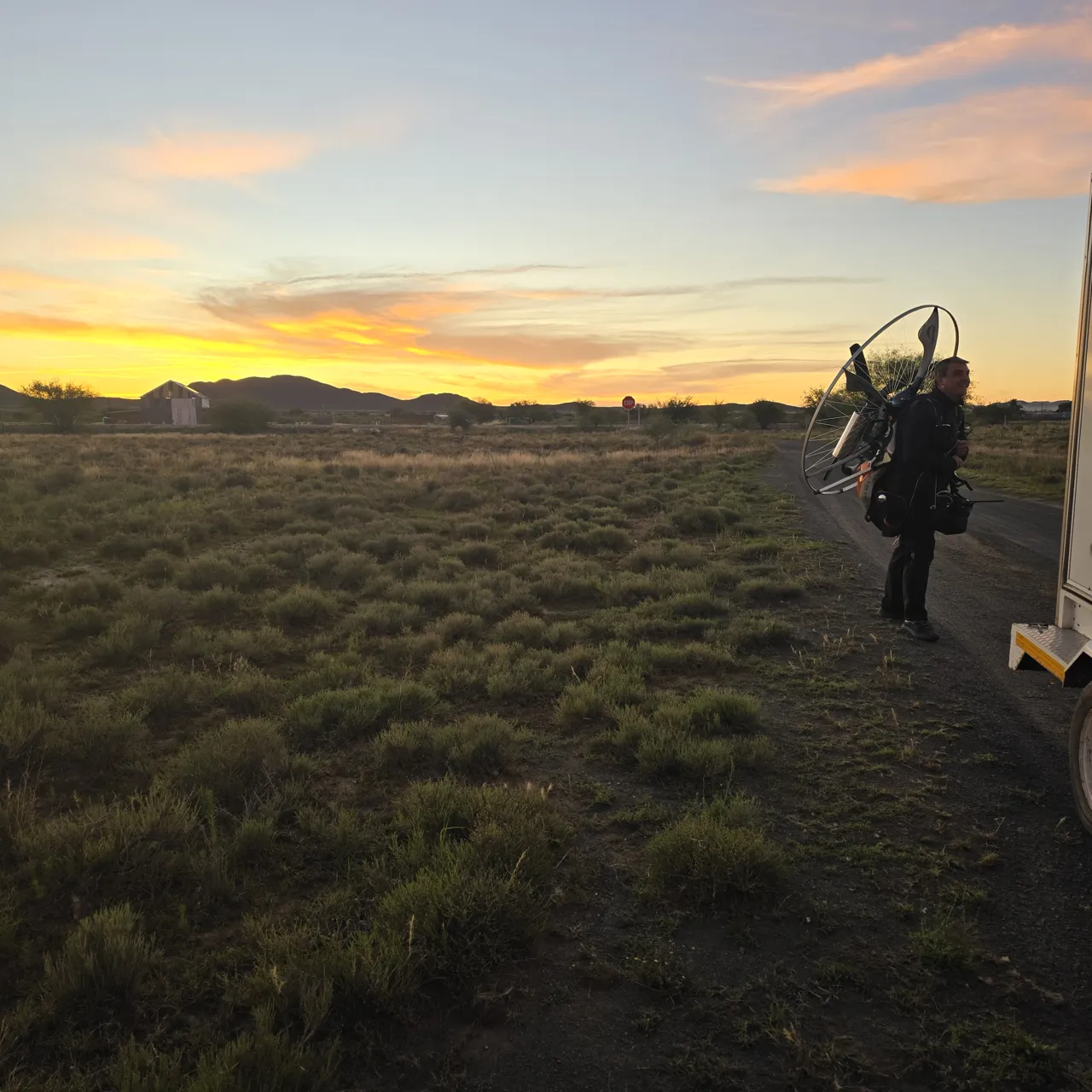 A pilot carrying a paramotor across grass at dusk on Day 2, with a trailer beside him