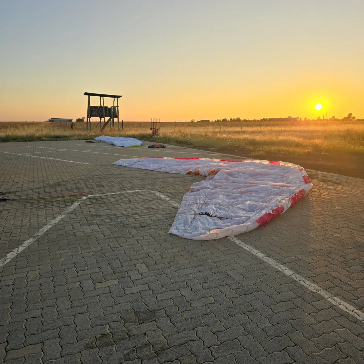 A paramotor wing laid out on paving at sunrise on Day 2, with a wooden lookout tower behind