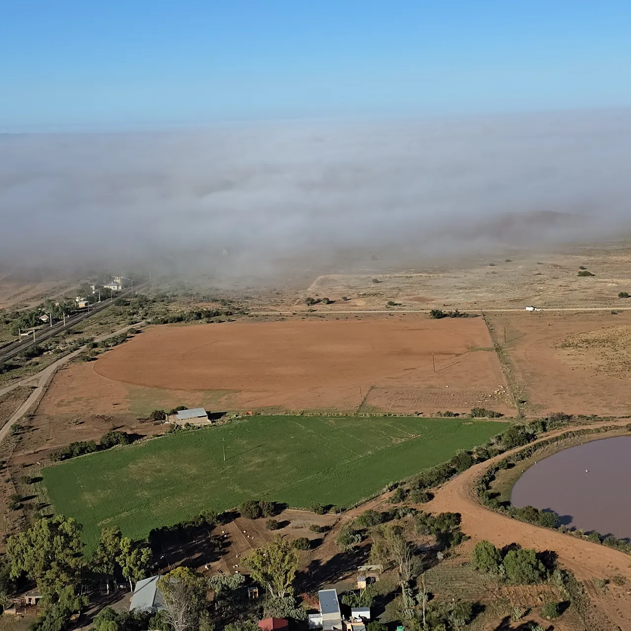 Low mist lying over farmland, with a green field and a dam, seen from the air on Day 3