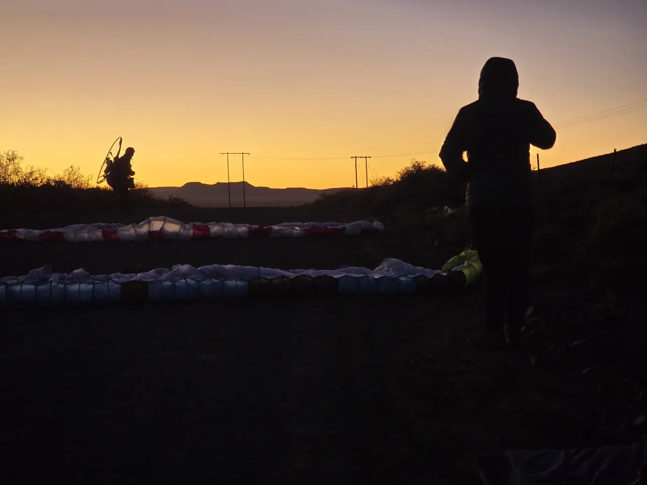 Two wings laid out on a dirt road at sunrise on Day 3, with a pilot in silhouette