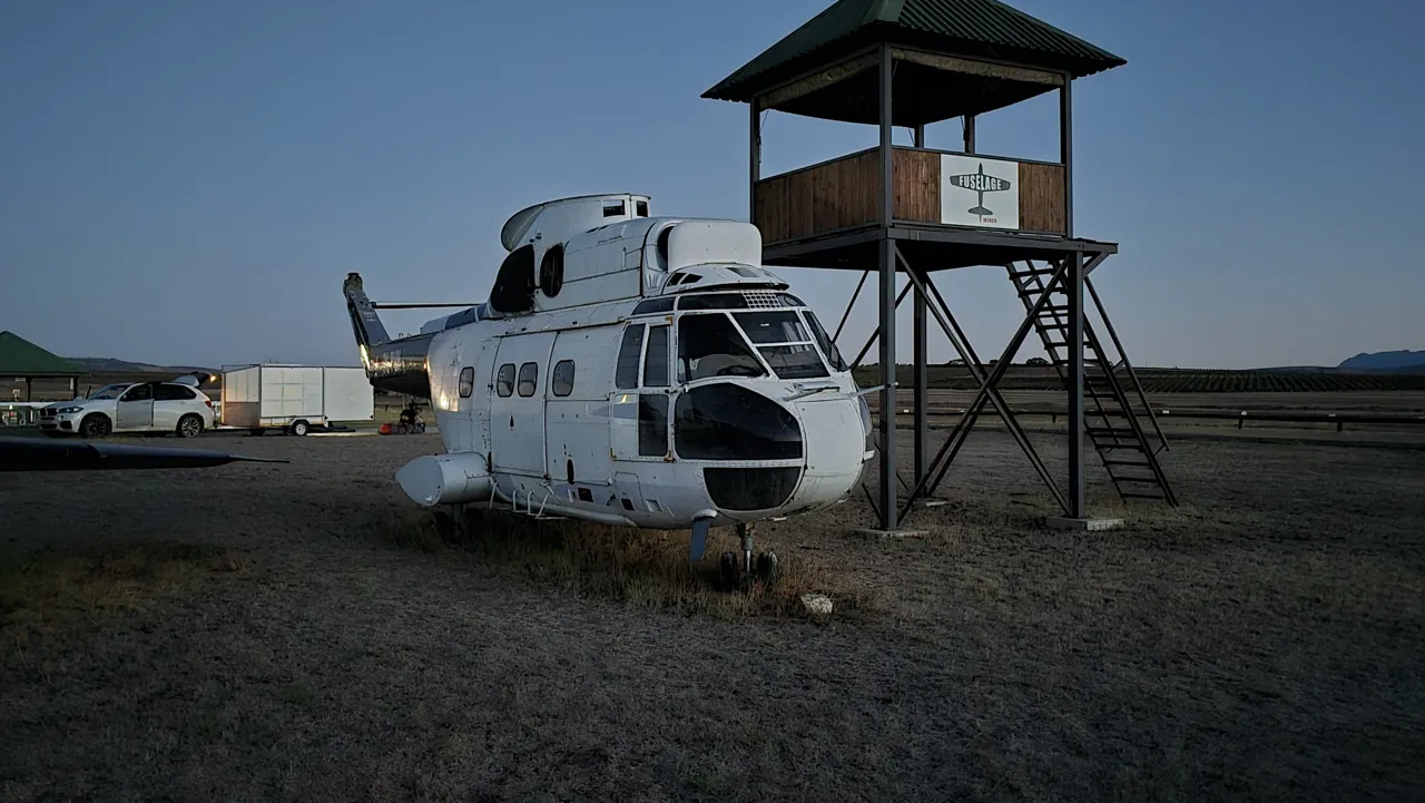 A helicopter on display beside a wooden lookout tower at Diemerskraal Airfield before dawn on Day 5