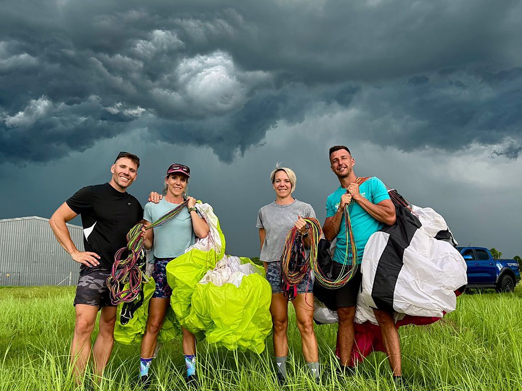Four pilots holding their wings and lines on grass under a dark storm sky after a flight