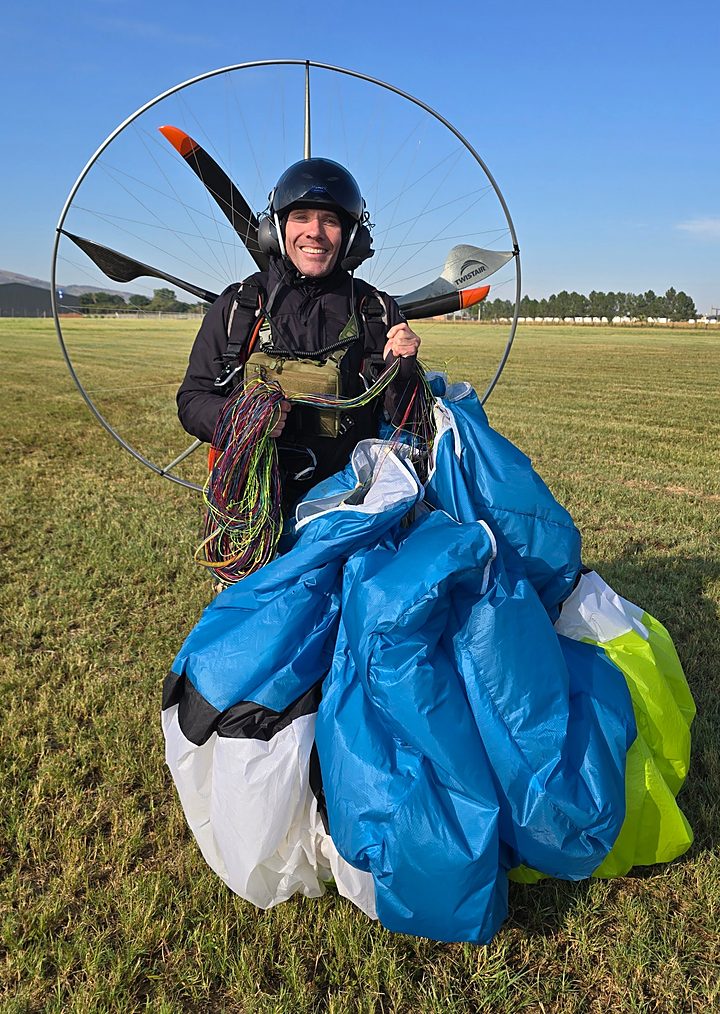 A smiling pilot holding a blue and lime wing after landing, paramotor frame on his back