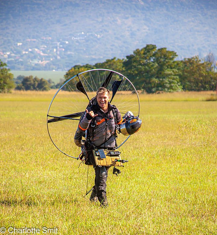 A smiling pilot walking across a grass field with a paramotor on his back
